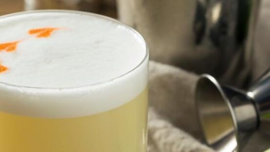 A frothy yellow cocktail in a glass with bitters on top, sitting near a cocktail shaker and a sliced lime.