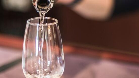 A person pours white wine from a bottle into a clear tasting glass on a dark wooden bar counter.
