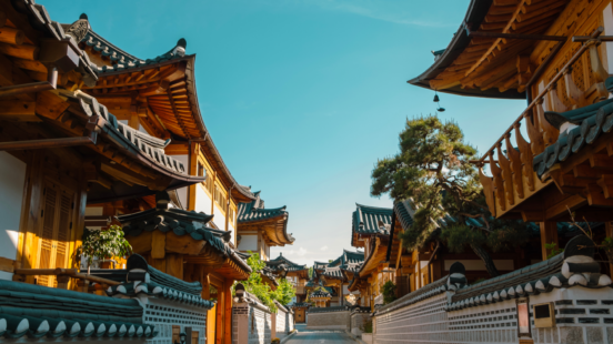 A narrow street lined with traditional Korean hanok houses featuring dark tiled roofs and light wooden frames under a blue sky.