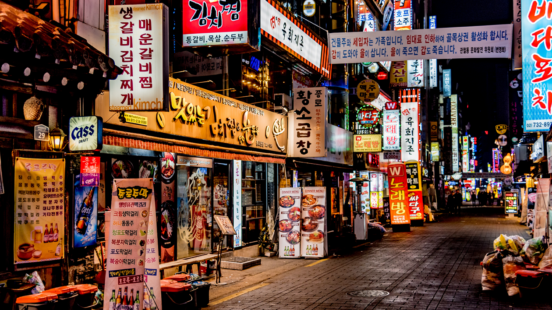 A narrow street in a South Korean city at night, lined with brightly lit buildings, numerous colorful neon signs, and restaurant storefronts.