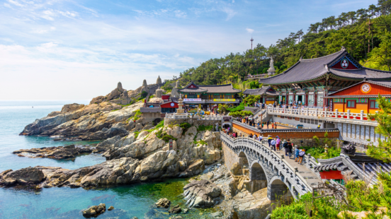 Haedong Yonggungsa Temple complex on a rugged coastline in Busan, South Korea, with a stone bridge and clear blue ocean.