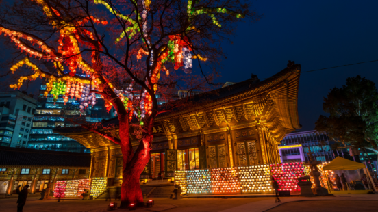 A Buddhist temple lit up at night with golden light and many colorful lanterns hanging on a large tree in the foreground.