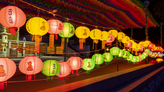 Strings of brightly colored paper lotus lanterns in pink, yellow, and green hanging in a row over a stone walkway at night.