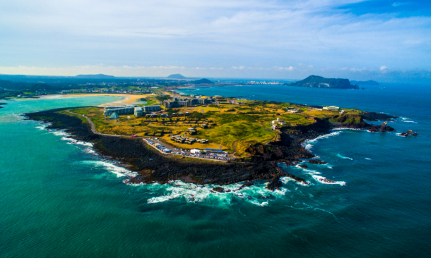 Aerial view of a resort complex on a rocky, grassy peninsula extending into the turquoise ocean, with coastline and hills visible in the background.