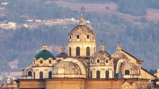 A view of a historic cathedral with several domes and towers against a backdrop of hazy blue hills.