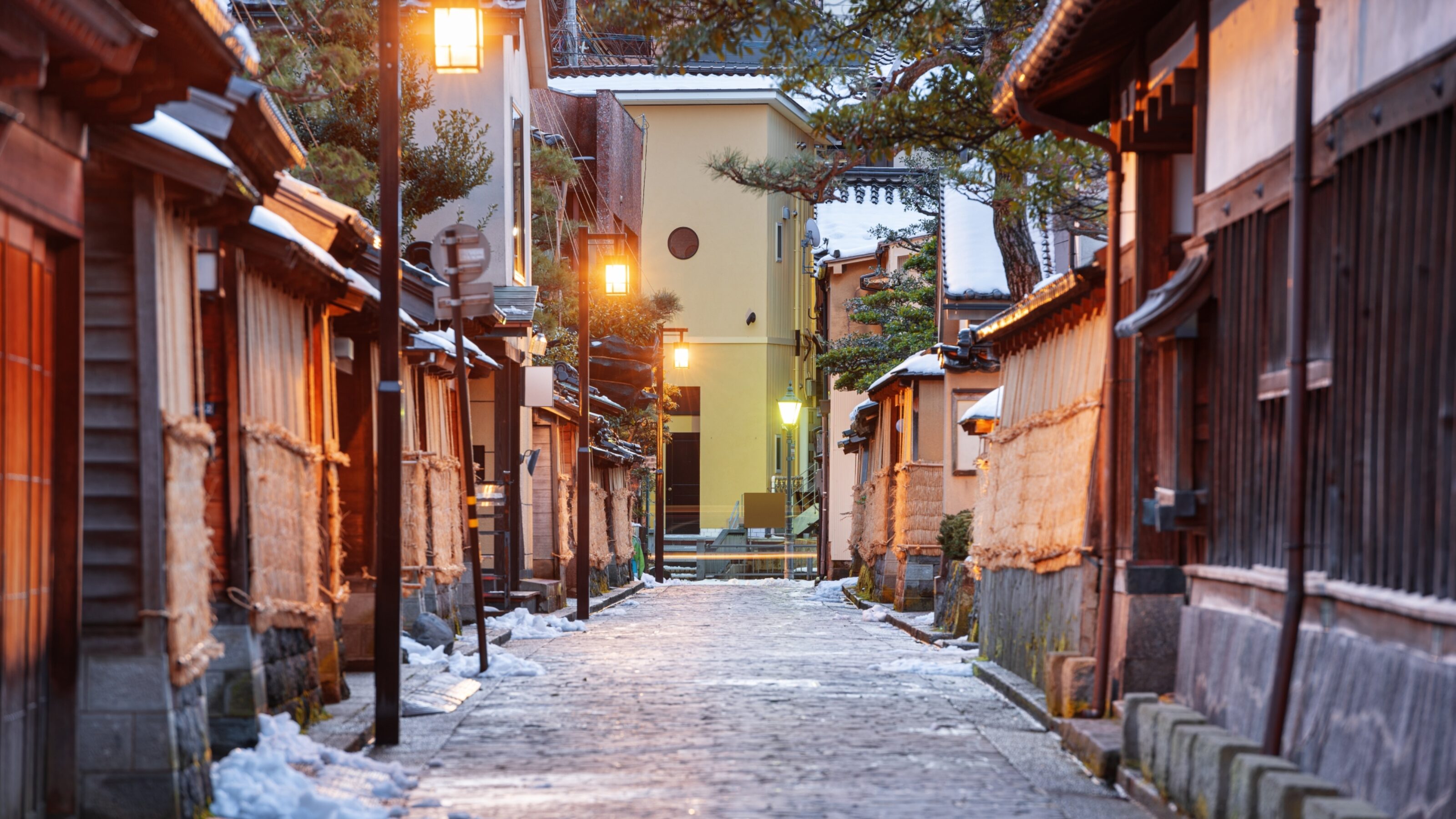 A quiet cobblestone street in Japan at dusk with glowing lanterns and patches of snow on the ground.