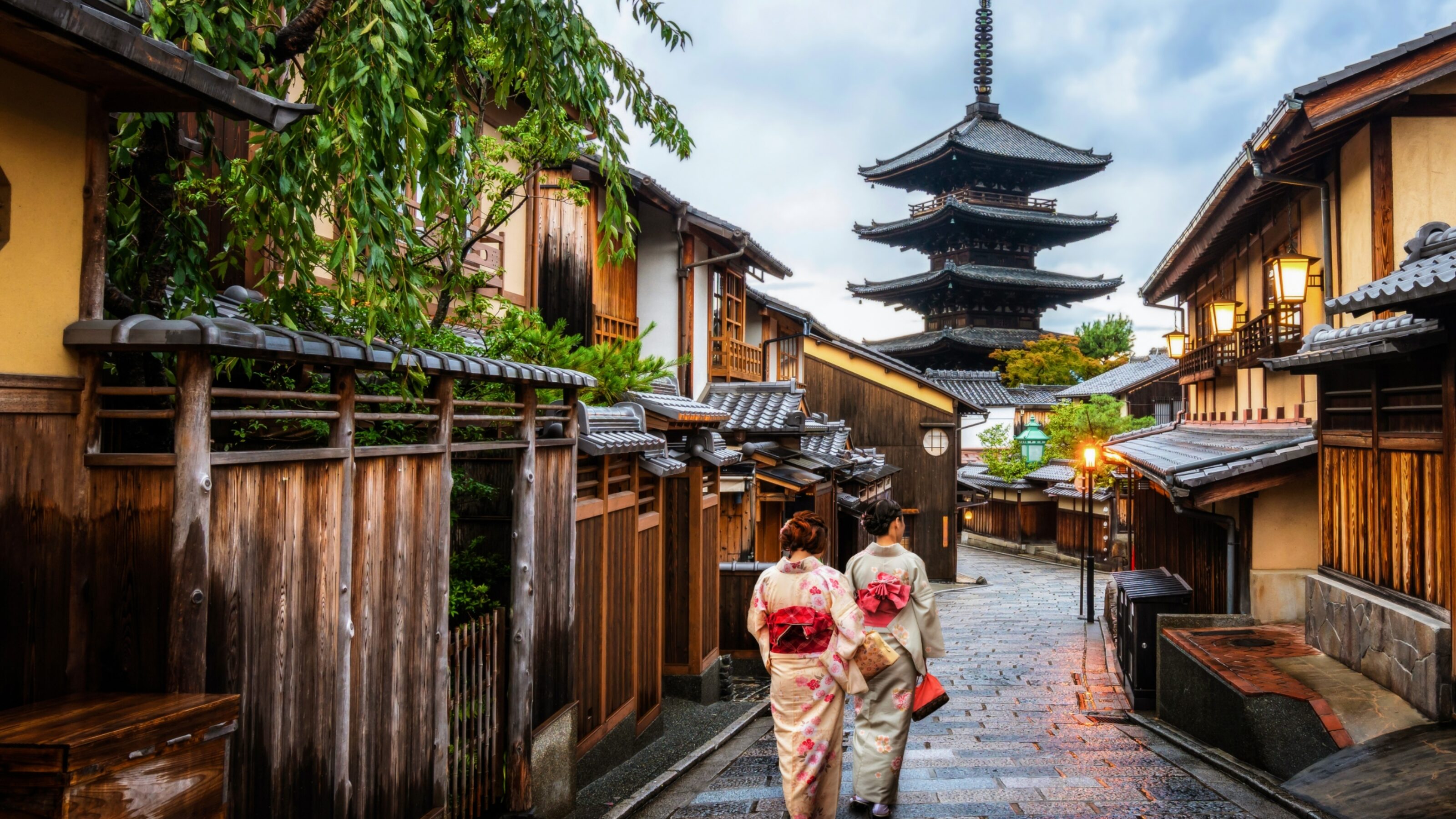 Two people in kimonos walk down a narrow Japanese street with wooden houses and a pagoda in the distance.
