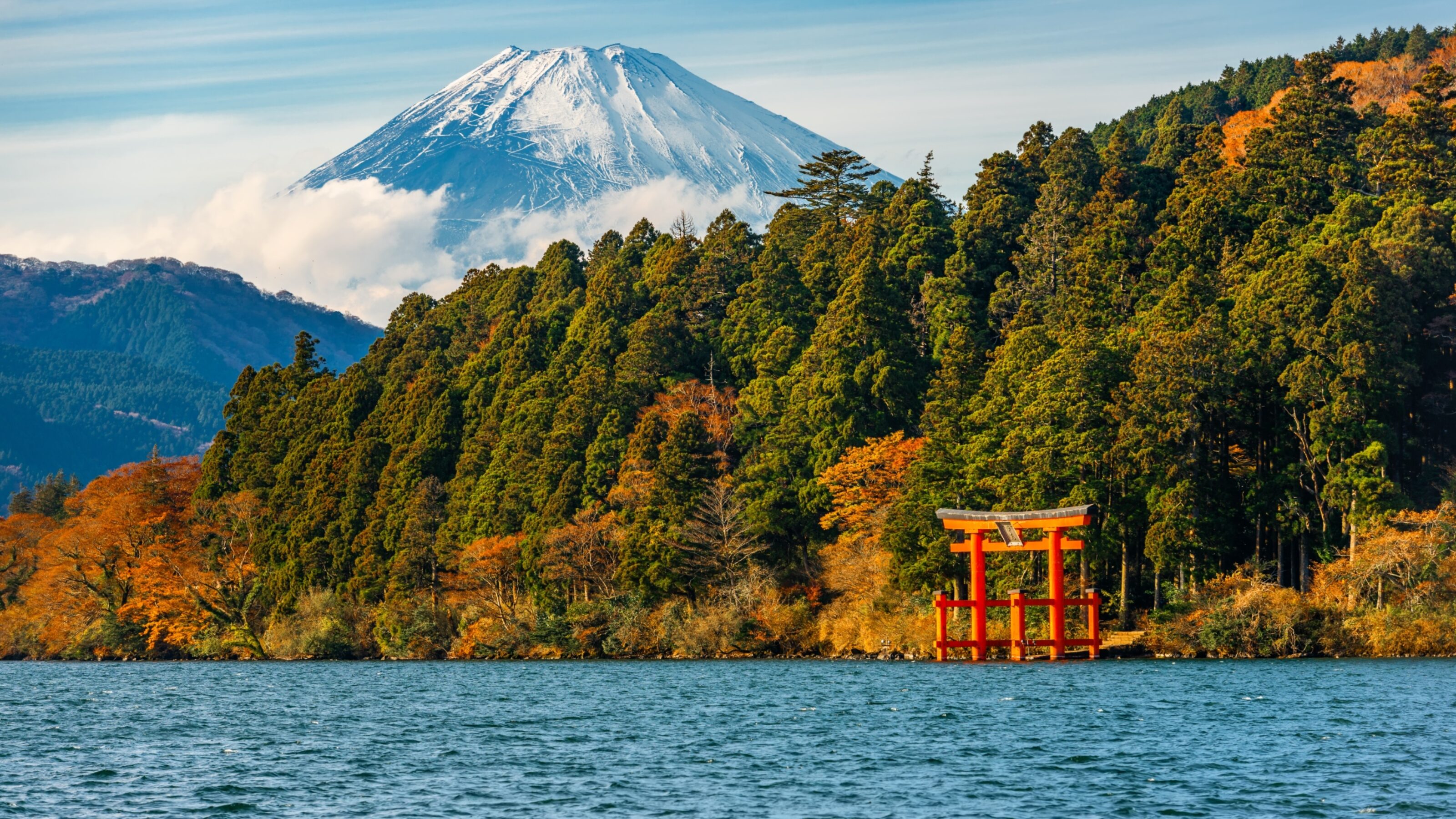 Snow-capped Mount Fuji behind a blue lake and a red torii gate by the forest shore.