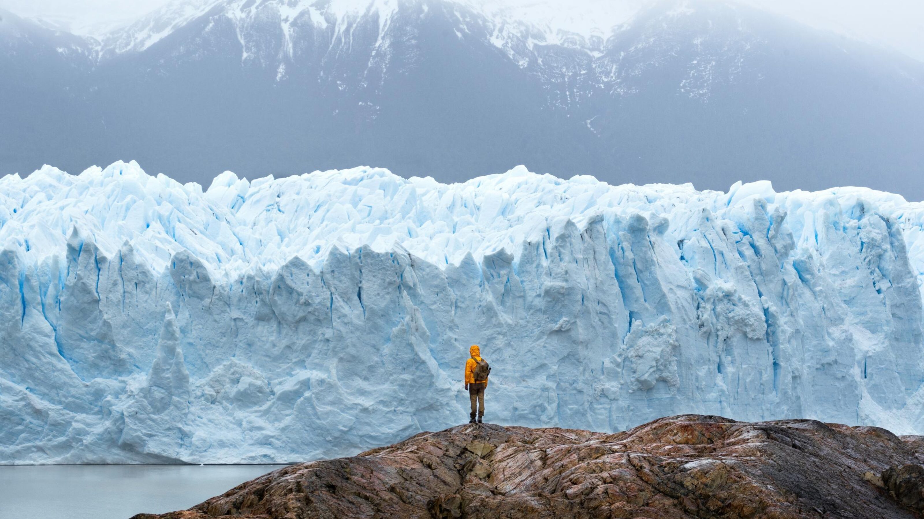 Person in yellow jacket standing before a massive glacier and mountains.