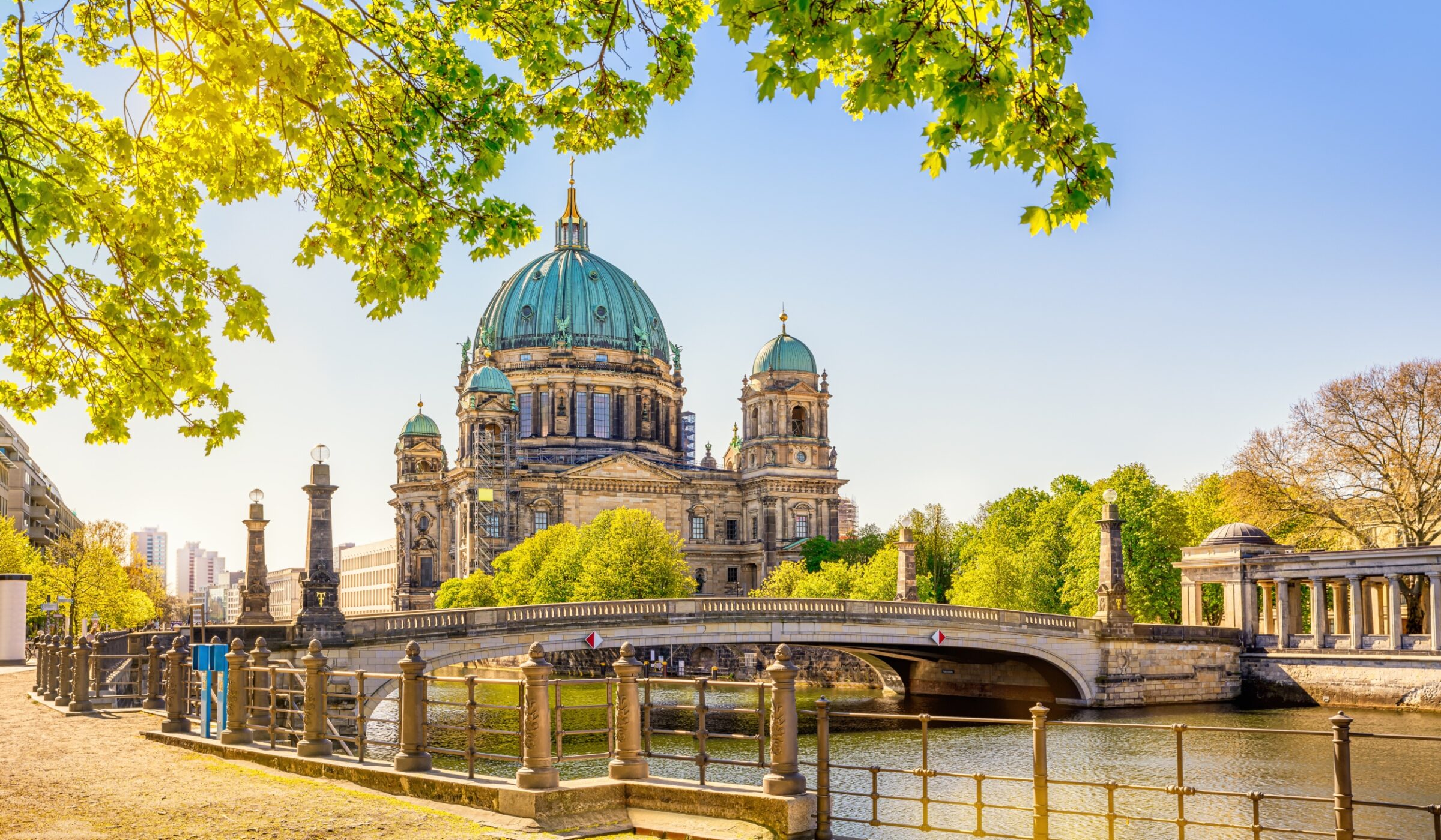 Berlin Cathedral with bridge over Spree River, framed by green foliage on a sunny day.