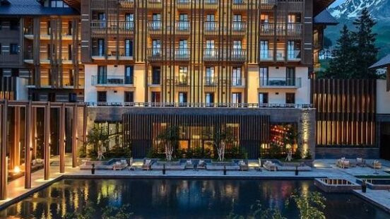 Luxury mountain resort with a pool at dusk, featuring a large wooden façade and seating area.