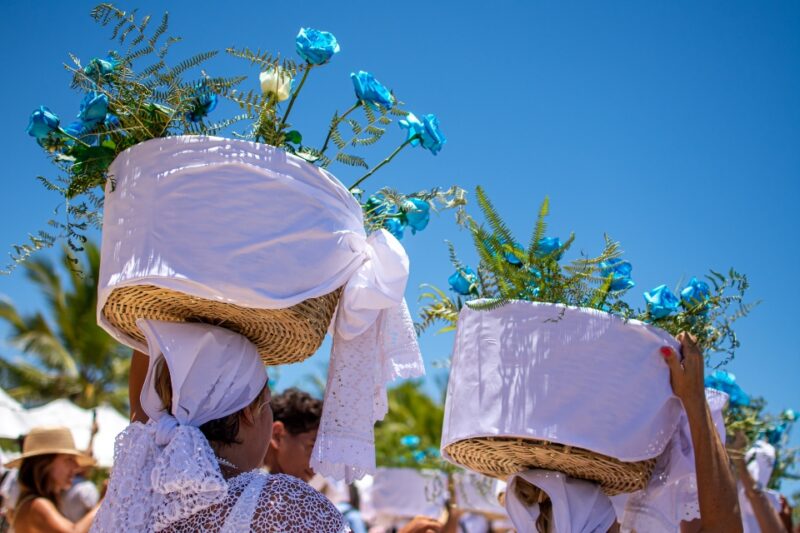 Two people wearing white headscarves and carrying baskets adorned with blue flowers on their heads.