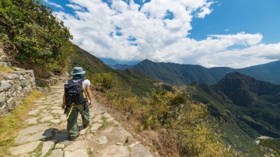 Hiker on mountain path with scenic valley view and cloudy sky.
