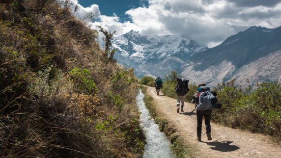 Hikers on a mountain trail with a stream alongside, snowy peaks in the background.