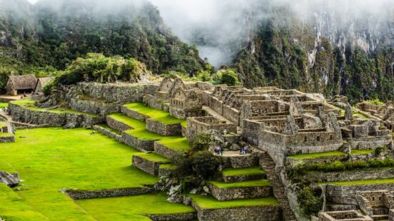 Ancient stone ruins of Machu Picchu with terraces, surrounded by greenery and fog-covered mountains.