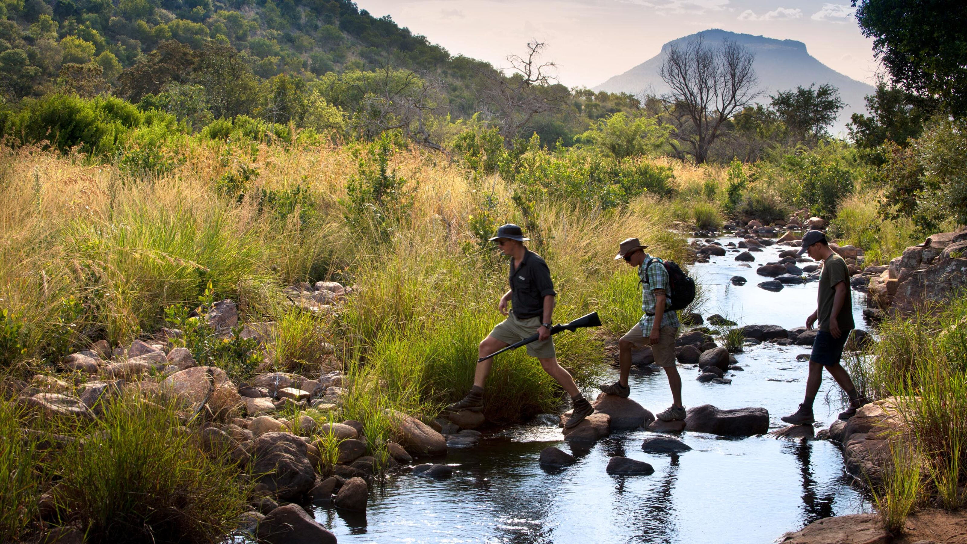 Three individuals crossing a rocky stream in a grassy landscape with a mountain backdrop.