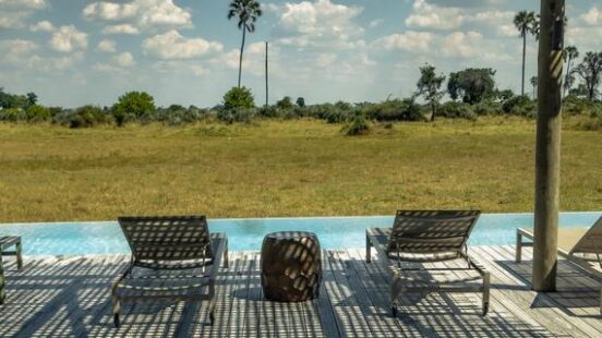 Poolside lounge chairs under a slatted roof with a view of a sunny savannah and palm trees.