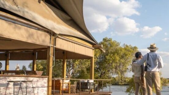 Two people standing by a safari tent lodge near a calm water body under a clear sky.