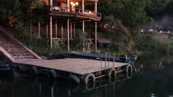 Two-story wooden cabin on stilts by a river at dusk, with lit interior and a dock.