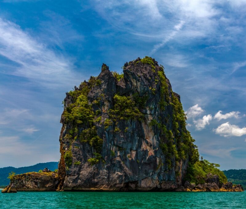Rocky island with lush greenery in turquoise sea under blue sky with clouds.