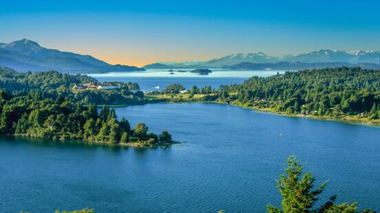 Lush greenery surrounds a serene lake with distant snow-capped mountains under a clear blue sky.