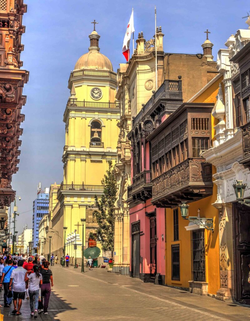 A bustling street scene with pedestrians, historic yellow tower with clock and Peruvian flag, and colonial balconies.