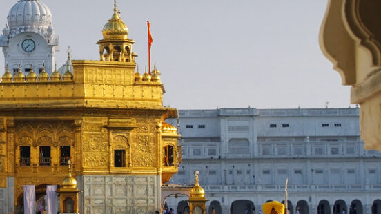 A person in traditional attire stands beside the reflective pool of the Golden Temple at sunrise.