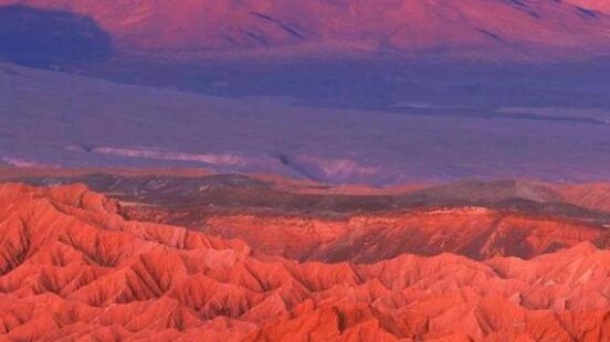 Reddish hues bathe a rugged landscape with a conical volcano in the background.
