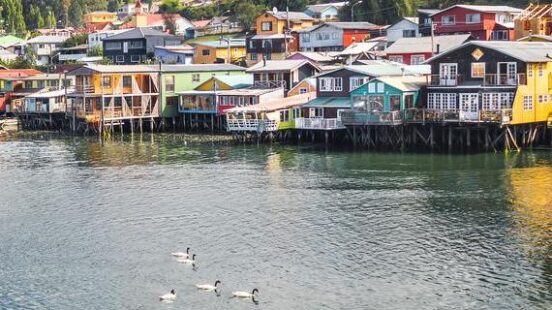 Colorful stilt houses by the water with a rowboat in the foreground and birds swimming.