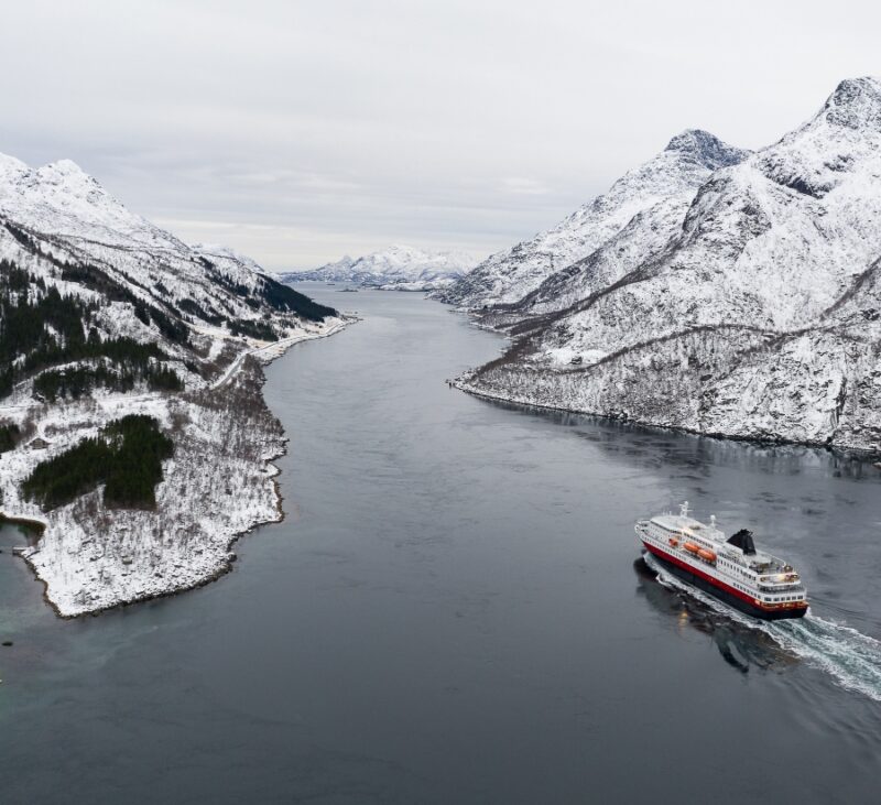 Aerial view of a cruise ship sailing through a snow-covered fjord surrounded by mountainous terrain.