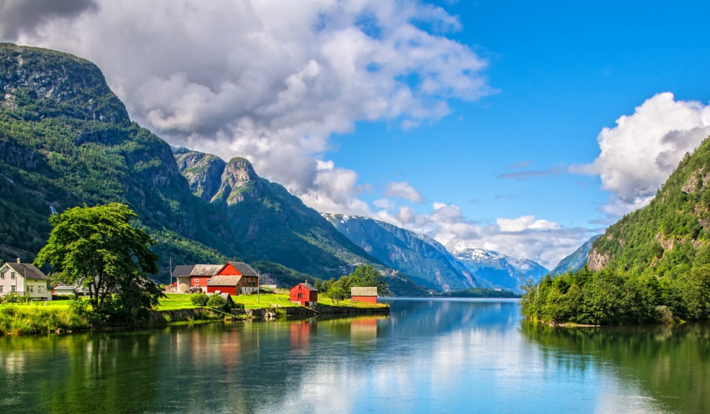 Red wooden cabins hug the shoreline along a Norwegian fjord – quiet waters reflecting steep mountain peaks.