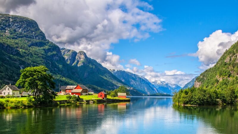 Red wooden cabins hug the shoreline along a Norwegian fjord – quiet waters reflecting steep mountain peaks.