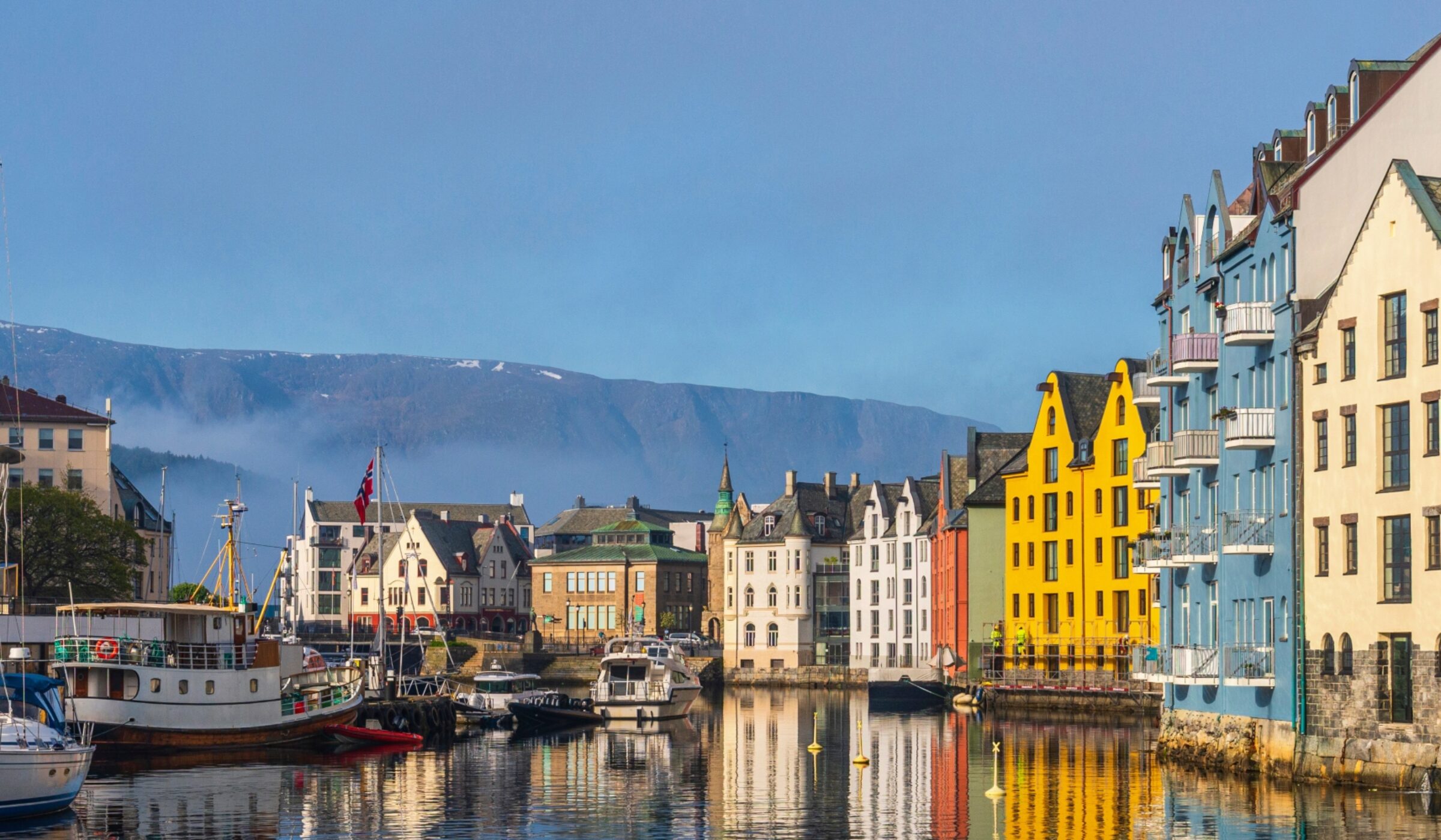 Colorful buildings by a calm harbor with boats, against a backdrop of mountains.