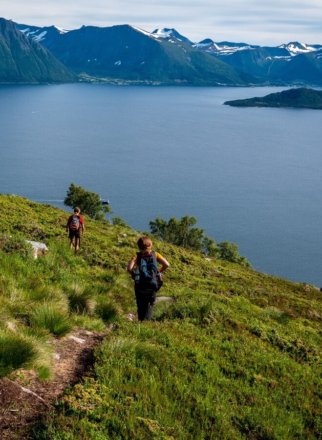 Two hikers trekking a hillside trail above a fjord with mountains in the background.