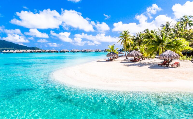 Tropical beach with clear blue water, white sand, palm trees, thatched umbrellas, and overwater bungalows.