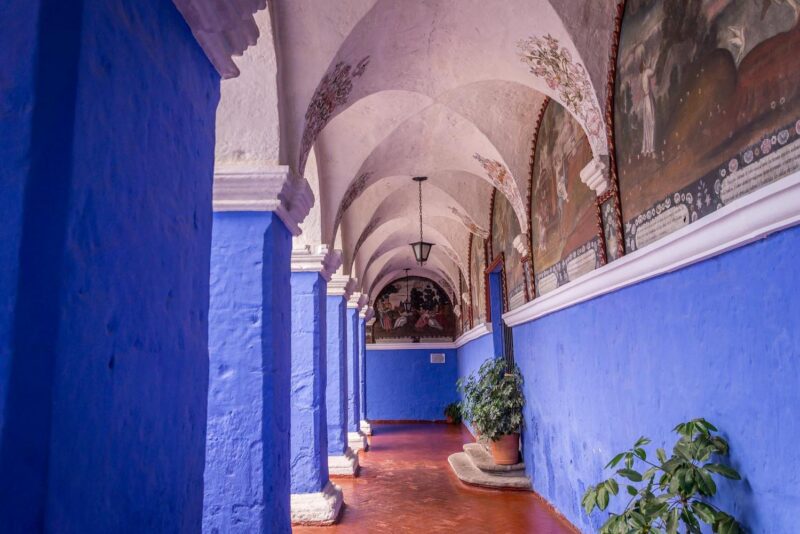 A vibrant blue hallway with arches, wall paintings, and potted plants.