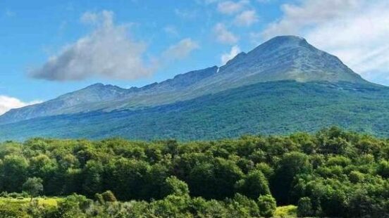 Panoramic view of Tierra del Fuego National Park, showing a volcano surrounded by green vegetation and water, against a blue sky.