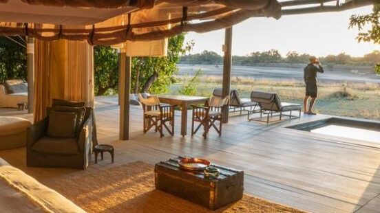 Luxury tent interior with open walls leading to an outdoor deck at sunset, person in distance enjoying the view.