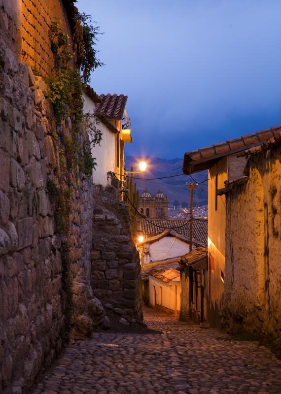 Narrow cobblestone street at twilight with stone walls and a distant church view.