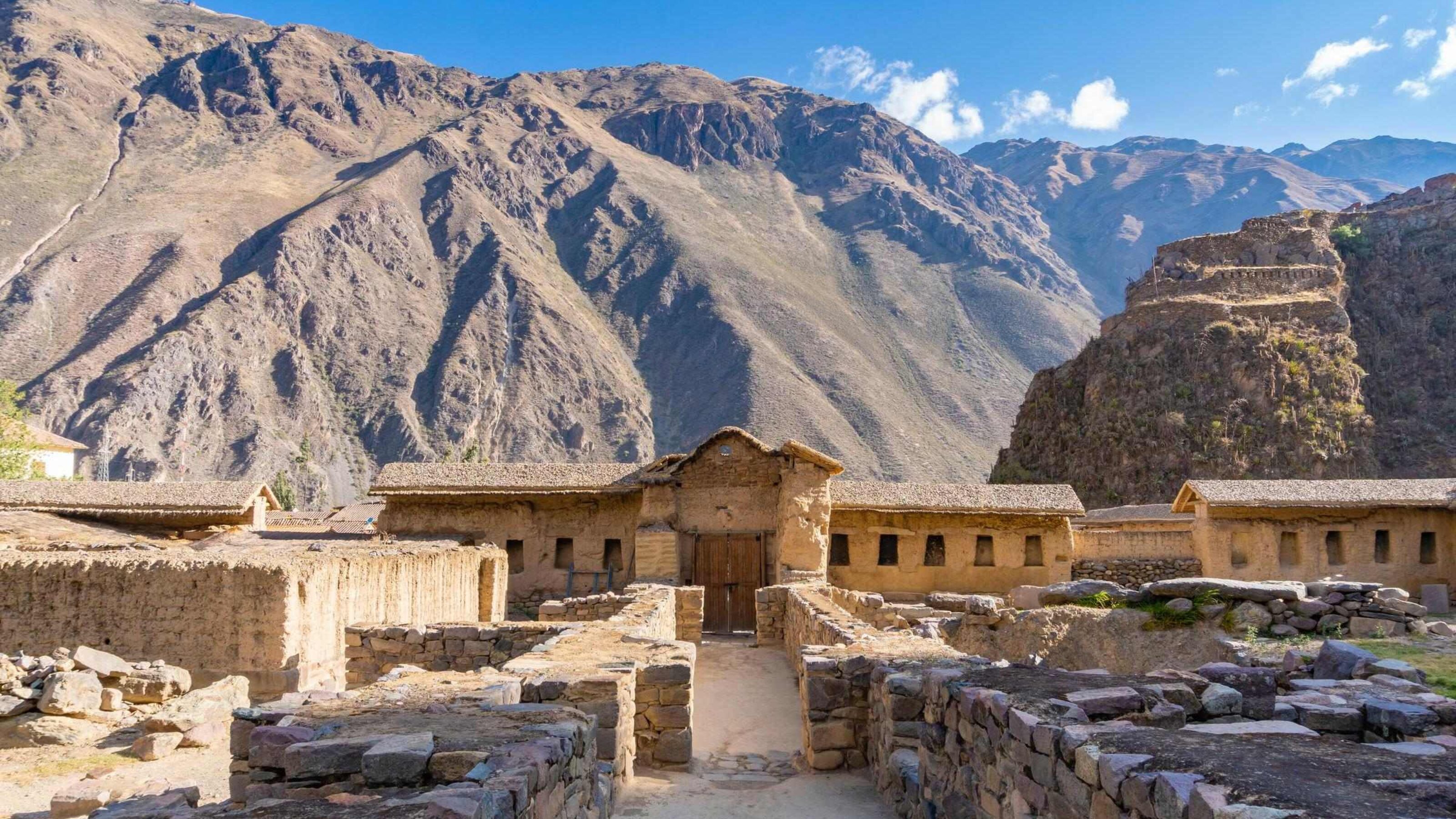 Ancient stone ruins with a thatched-roof hut against a backdrop of rugged mountains under a blue sky.