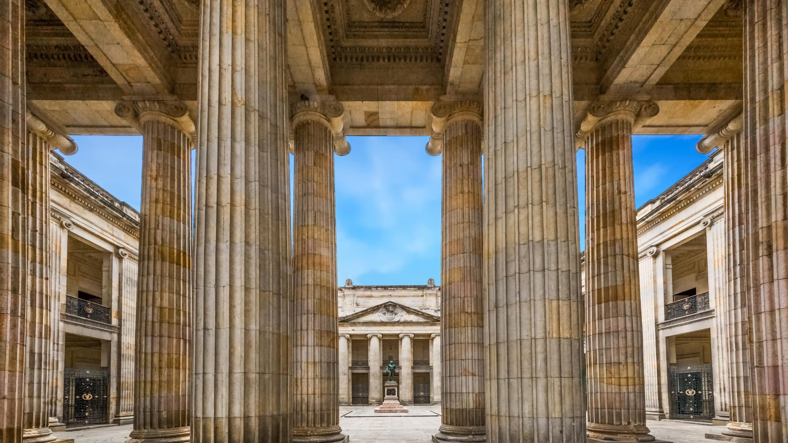 View through tall neoclassical columns to a courtyard with a statue and blue sky above.