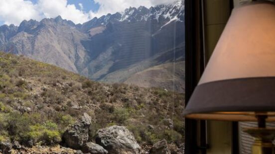 A view from a train window looks out at a rushing river and steep, rocky, scrub-covered mountainside with snow-capped peaks, a great luxury Machu Picchu holidays.