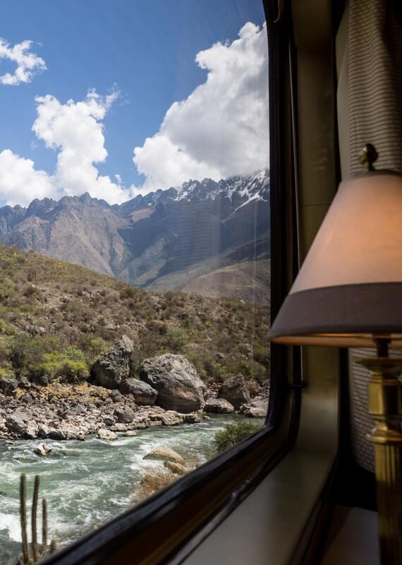 A view from a train window looks out at a rushing river and steep, rocky, scrub-covered mountainside with snow-capped peaks, a great luxury Machu Picchu holidays.