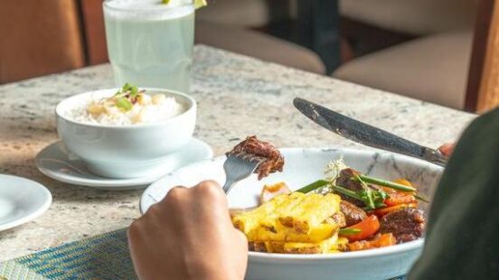 Person dining, with grilled vegetables and meat on a plate, a side of rice, and a lemonade.