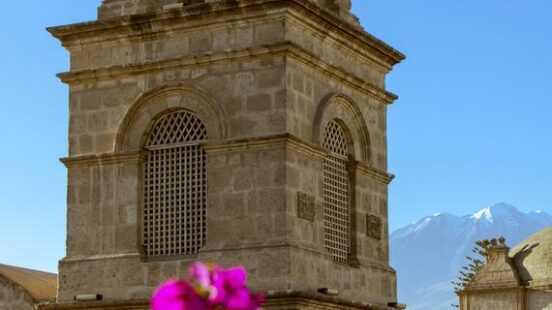 Stone church tower with a cross, framed by bright pink flowers, against a backdrop of snowy mountains.