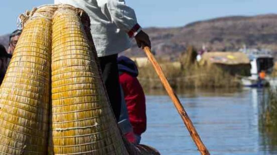 Person in traditional attire standing on a reed boat using a wooden pole to navigate the water.