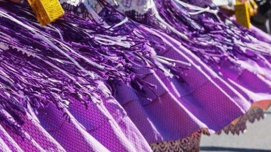 A row of individuals in vibrant purple traditional dresses and white hats carrying yellow ornaments.