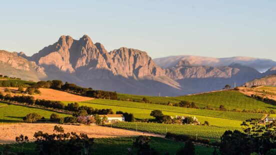 A scenic landscape of lush green fields and houses in front of large, rocky mountains.