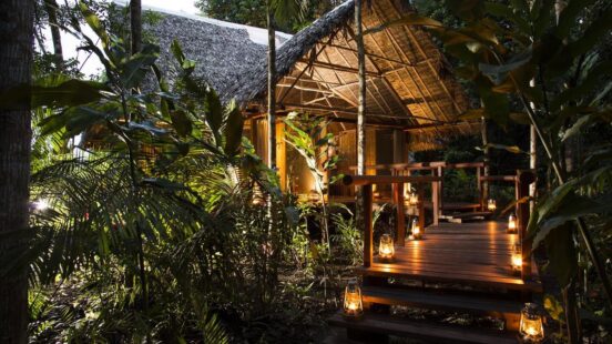 Thatched-roof hut illuminated by lanterns on a wooden deck surrounded by lush foliage at dusk.