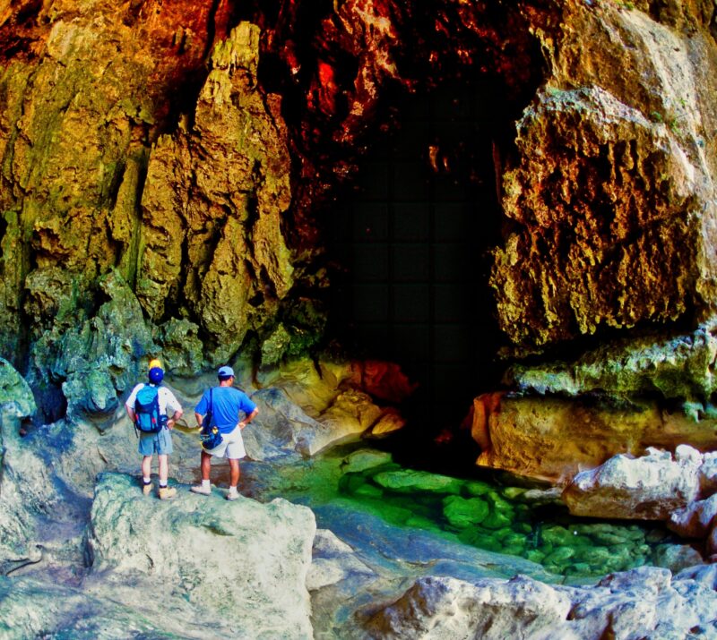 Two hikers with backpacks standing by a cave entrance surrounded by colorful rocks.
